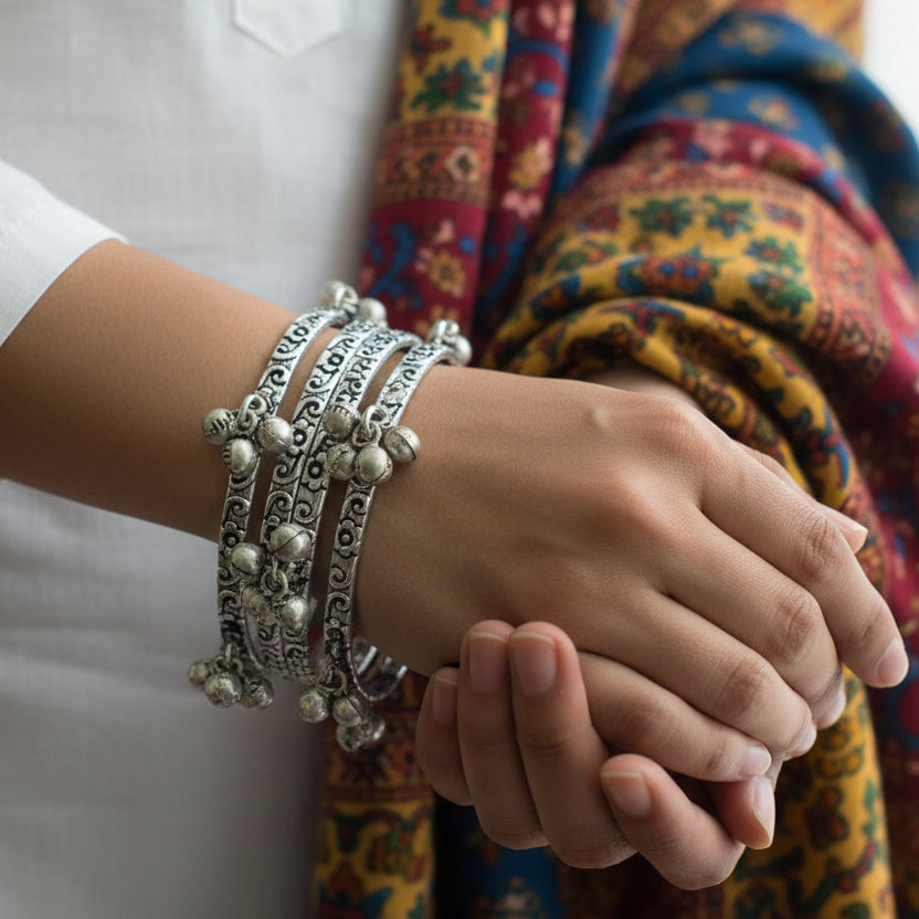 Silver bangles with intricate designs on a gray stand, branded 'Saheli'.
