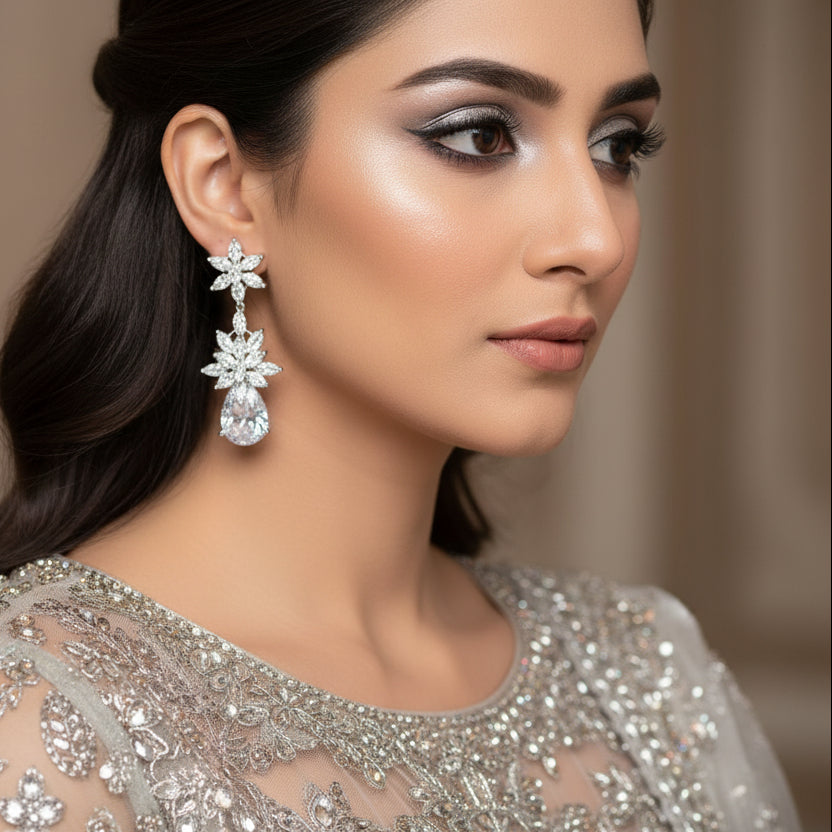 Pair of silver earrings with clear stones on a gray stand, branded 'Saheli'.
