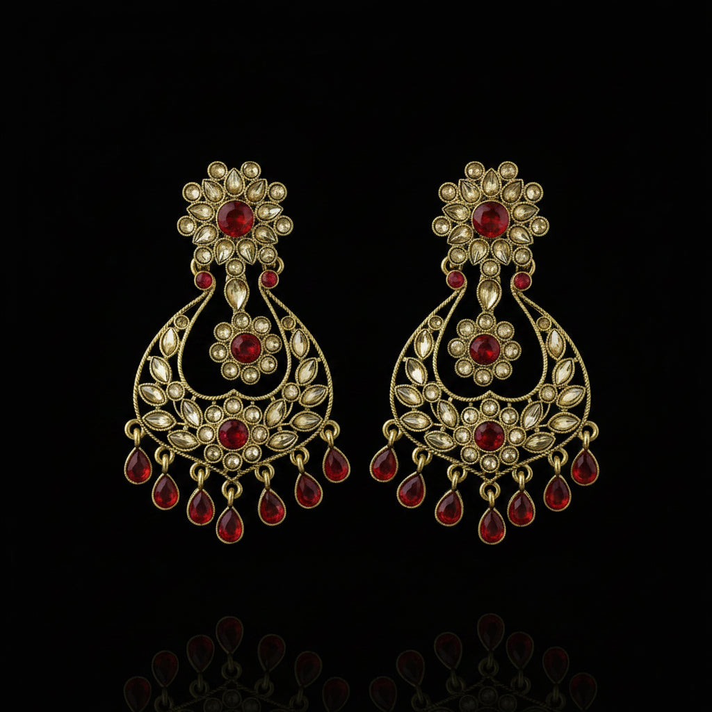 A pair of traditional Indian gold-plated chandelier earrings decorated with red stones and white stones, displayed on a dark gray background with a Saheli brand logo in the top left corner.