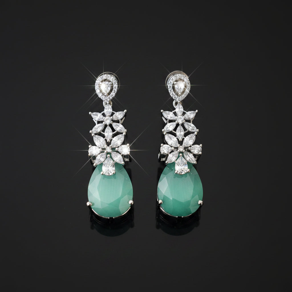 A pair of silver-tone drop earrings featuring a cluster of clear stones in a floral pattern above a teardrop-shaped light green jadeite stone, displayed on a dark reflective surface.