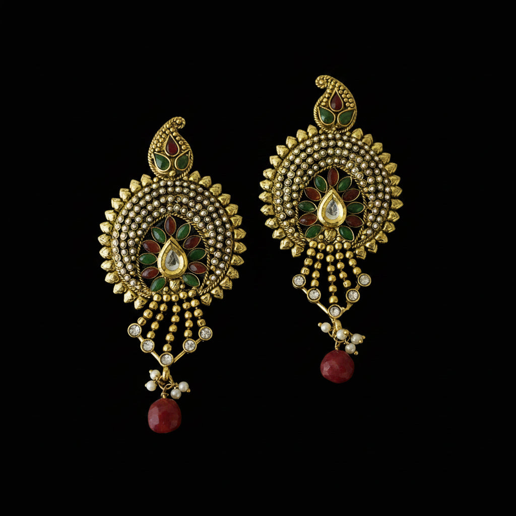 A pair of gold-plated Indian chandelier earrings with intricate green and red enamel work, white stone accents, and dangling red bead drops, displayed on a black reflective surface. The brand logo 'SAHELLI HARESTHIBH' is visible in the top left corner.