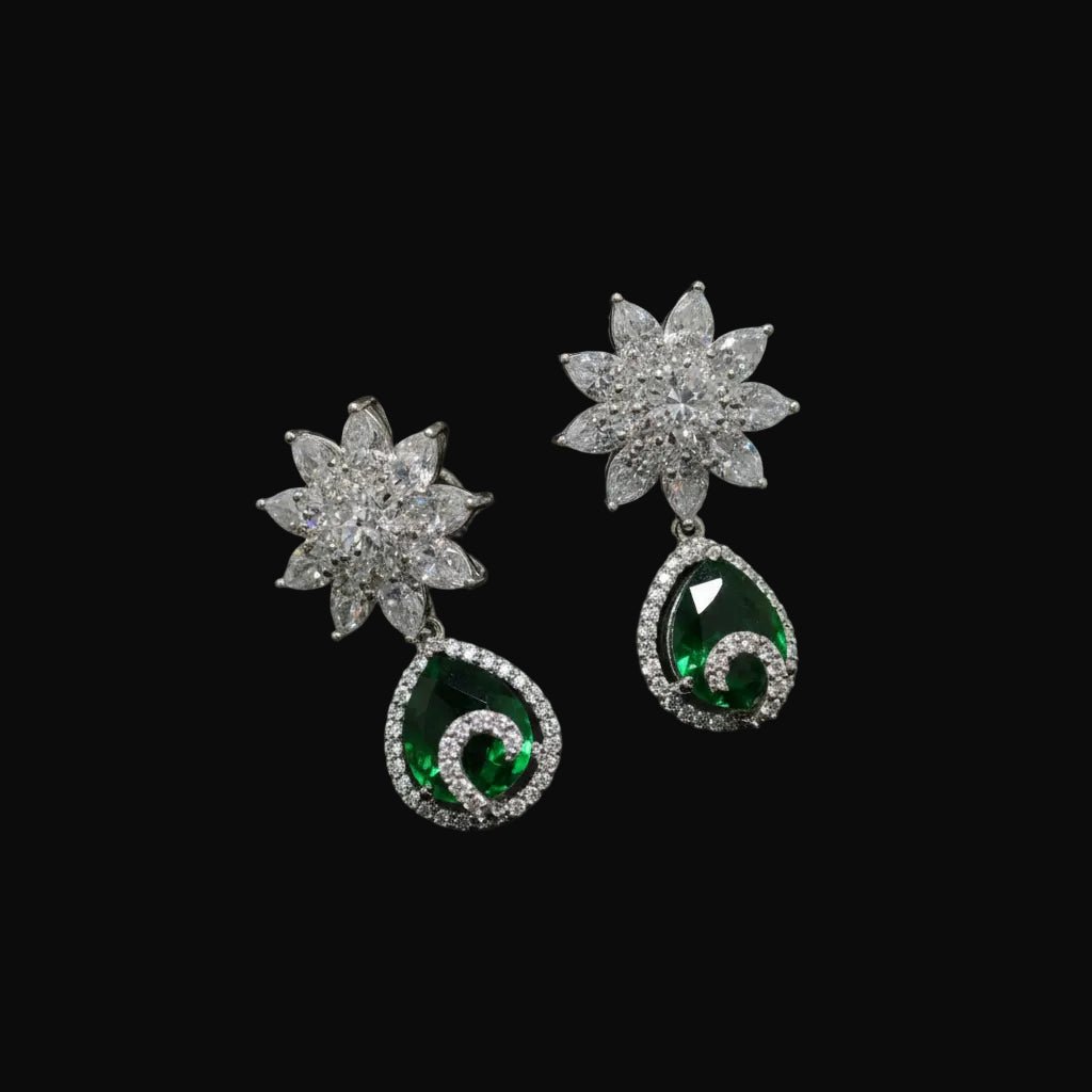 A pair of silver-toned dangle earrings with starburst-shaped clear stone tops and teardrop-shaped green stone drops, displayed on a dark textured fabric background. The brand name 'SAHELL' is visible at the top center.