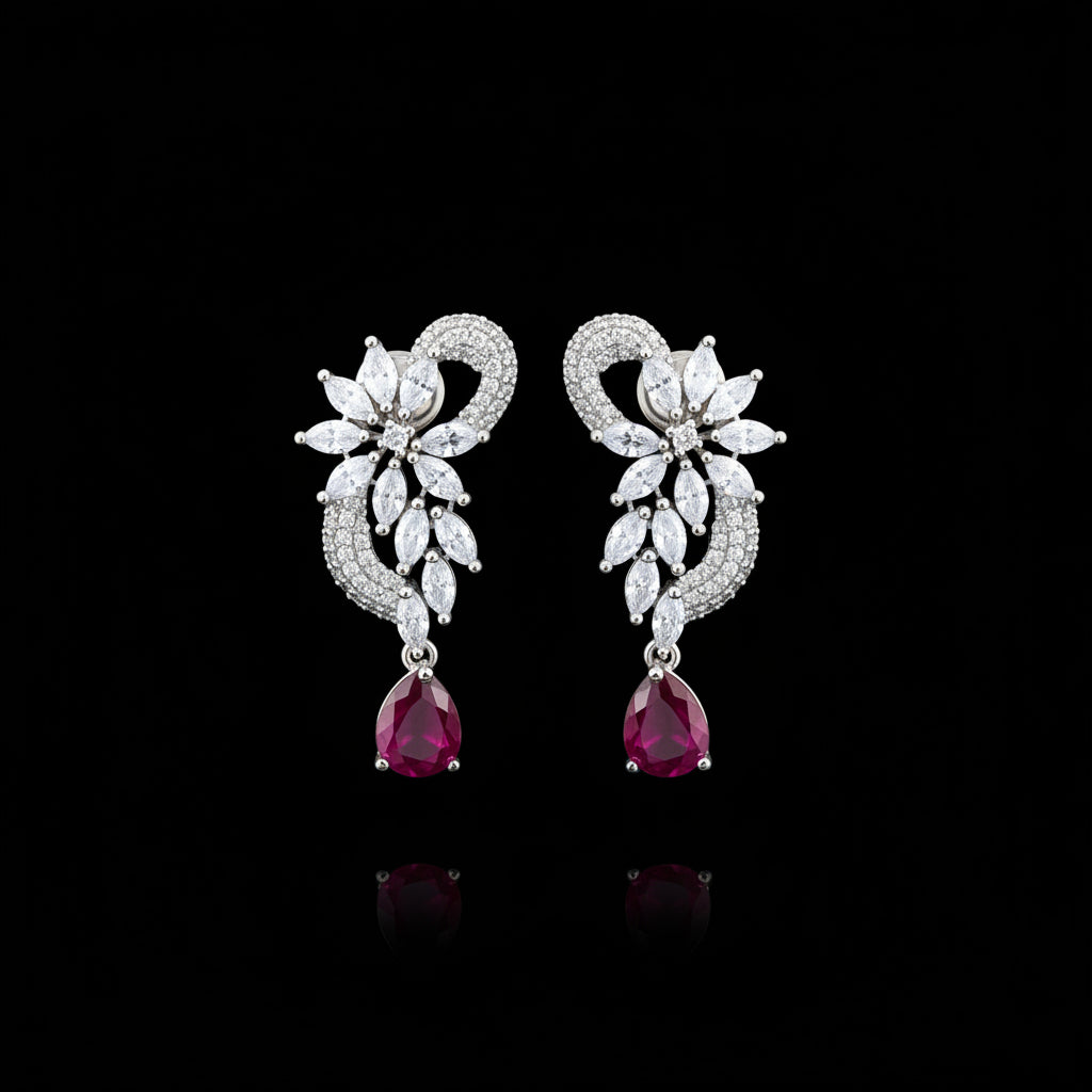 A pair of silver-toned statement earrings with clear pavé floral motifs and dangling teardrop-shaped red gemstones, displayed on a black stand against a gray background with the brand name SAHELI MY BEST FRIEND at the top.