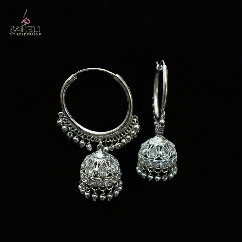 Silver hoop earrings with jhumka designs on a black stand against a dark background.