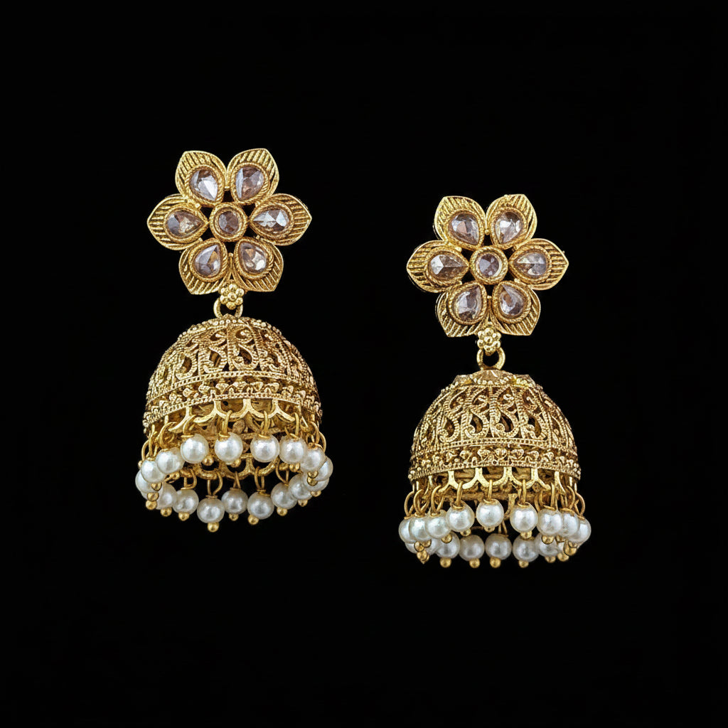 A pair of gold-plated Indian jhumka earrings with floral stud tops, clear rhinestones, and dangling pearl bead fringes, displayed on a solid dark purple background.