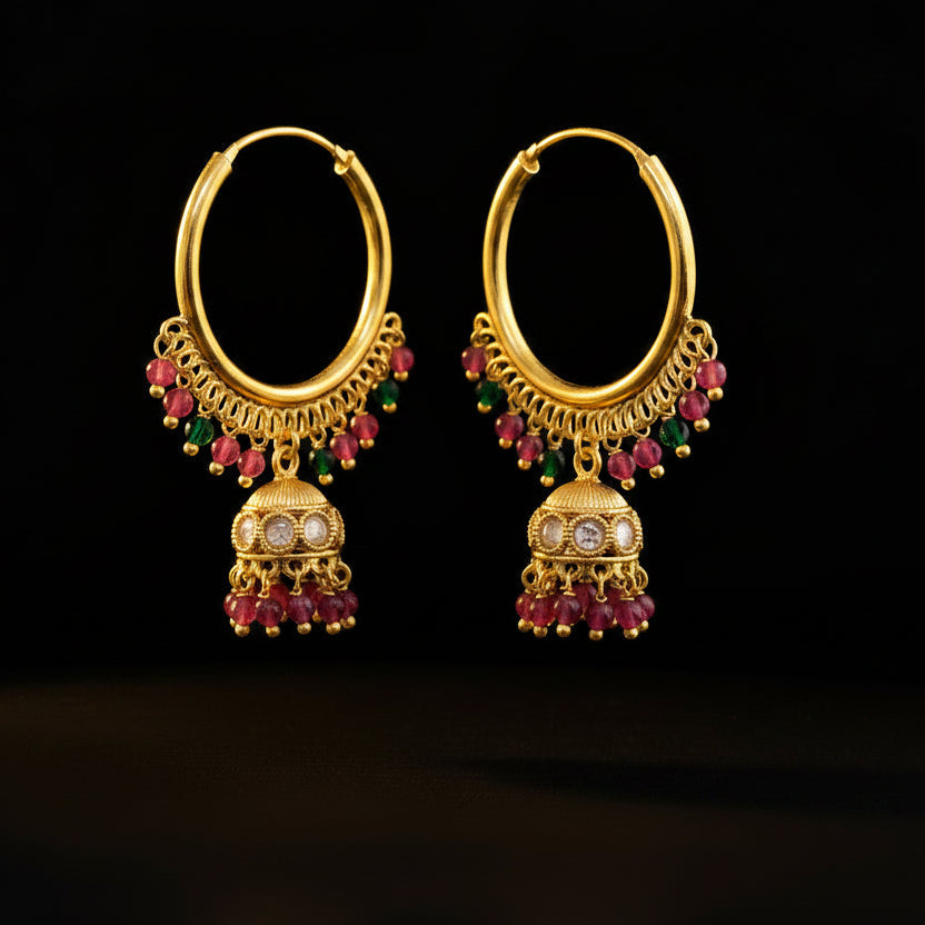 A pair of gold-toned traditional hoop earrings with dangling jhumka-style charms, decorated with pink and green beads and small clear stones, displayed on a gray jewelry stand against a gray background. The brand name 'SAHELI MY BEST FRIEND' is printed at the top.