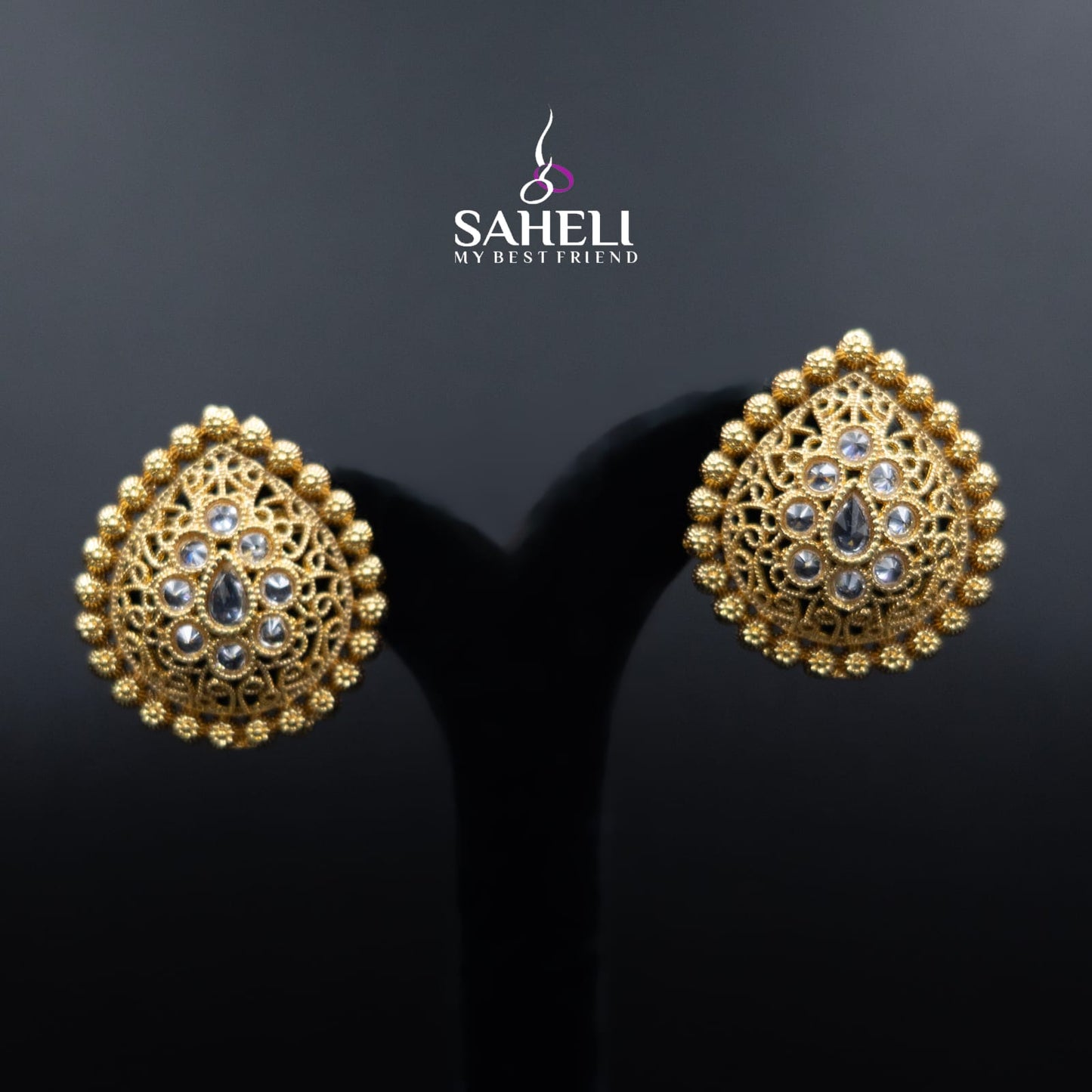 Gold earrings with intricate designs on a dark background, featuring the brand 'Saheli'.