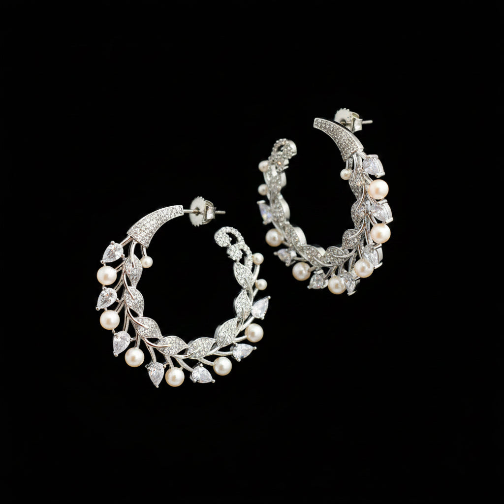 A pair of silver-toned crescent-shaped hoop earrings decorated with leaf and pearl motifs, displayed on a dark reflective surface.