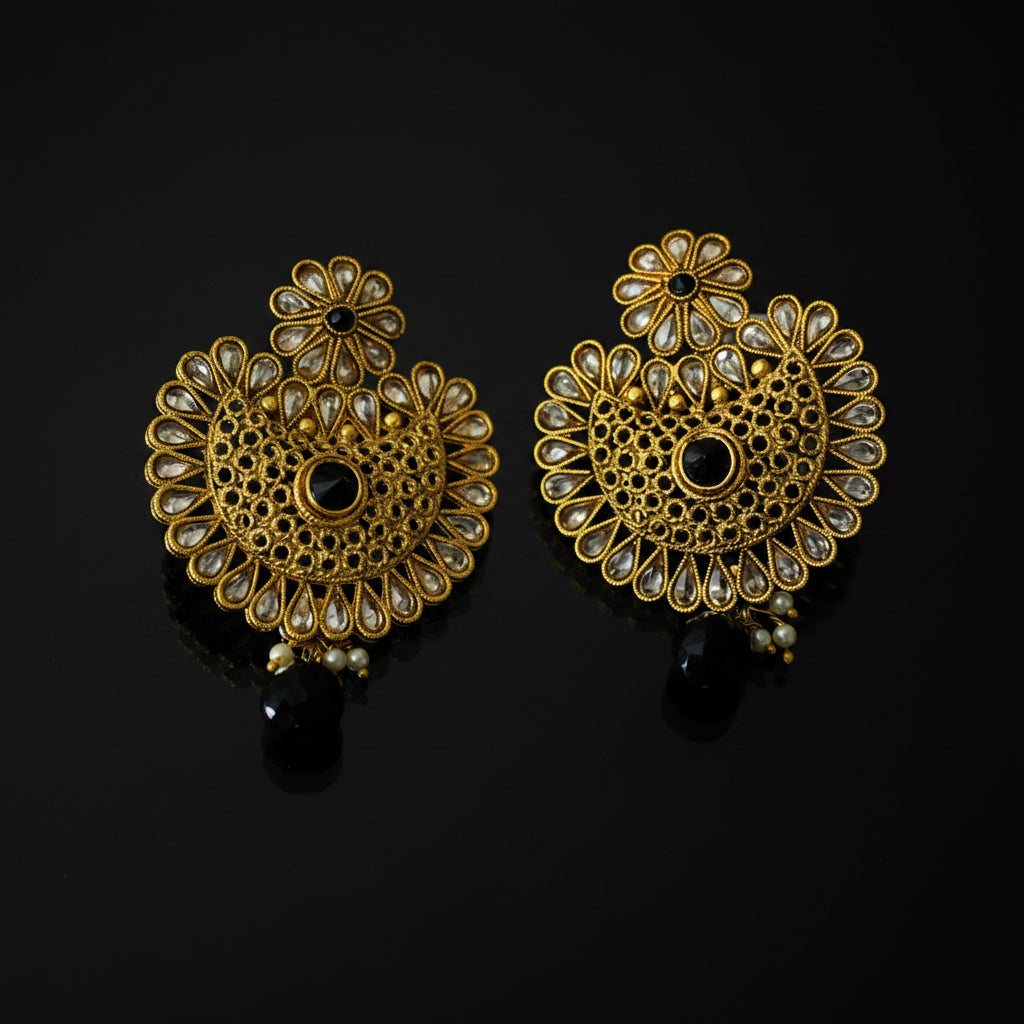 A pair of ornate gold-toned Indian jhumka earrings decorated with clear kundan stones and black bead accents, displayed on a reflective black surface. The brand logo 'SAHELI MY BESTFRIEND' is visible in the top left corner.