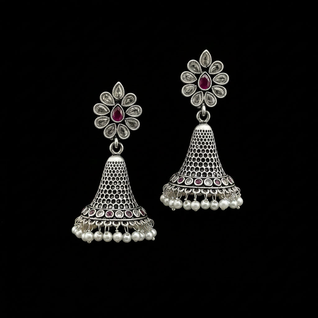 A pair of silver-toned Indian jhumka earrings with floral stud tops, pink stone accents, and dangling bell-shaped charms decorated with small white pearls, displayed on a gray stand.
