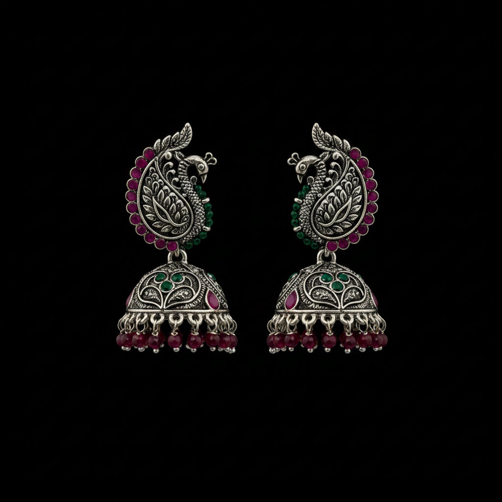 A pair of ornate silver-toned Indian jhumka earrings with peacock motifs, pink and green stone accents, and dangling bead chains, displayed on a black jewelry stand against a dark gray background.