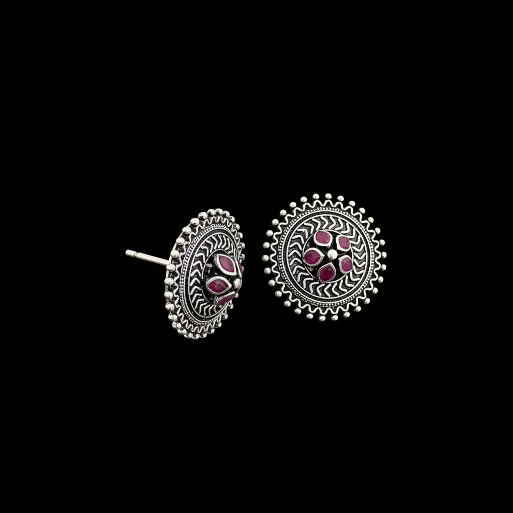 A pair of round silver-toned earrings with intricate filigree patterns and small pink gemstone accents, displayed on a dark reflective surface.