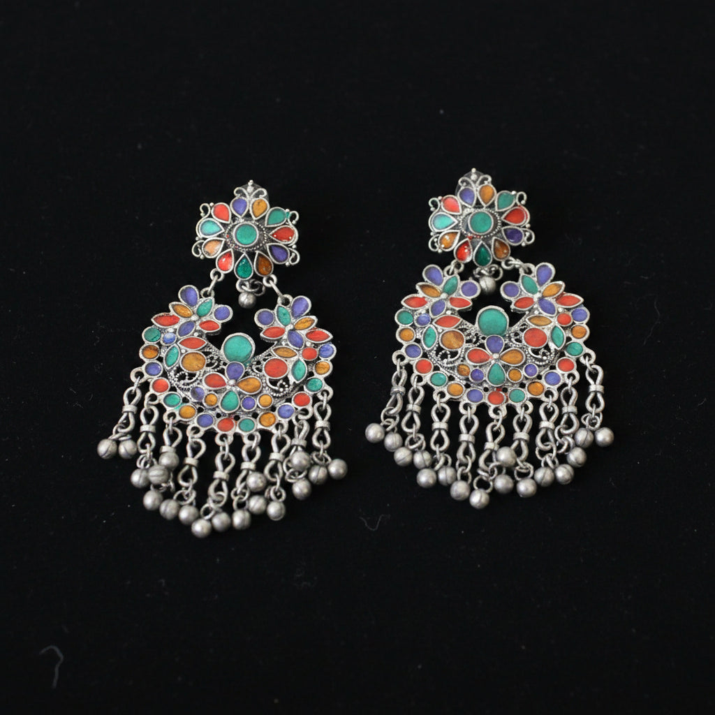 A pair of ornate silver-toned Indian chandbali earrings decorated with multicolored enamel in green, orange, red, and purple, featuring dangling silver chains and bead clusters on a dark reflective surface.