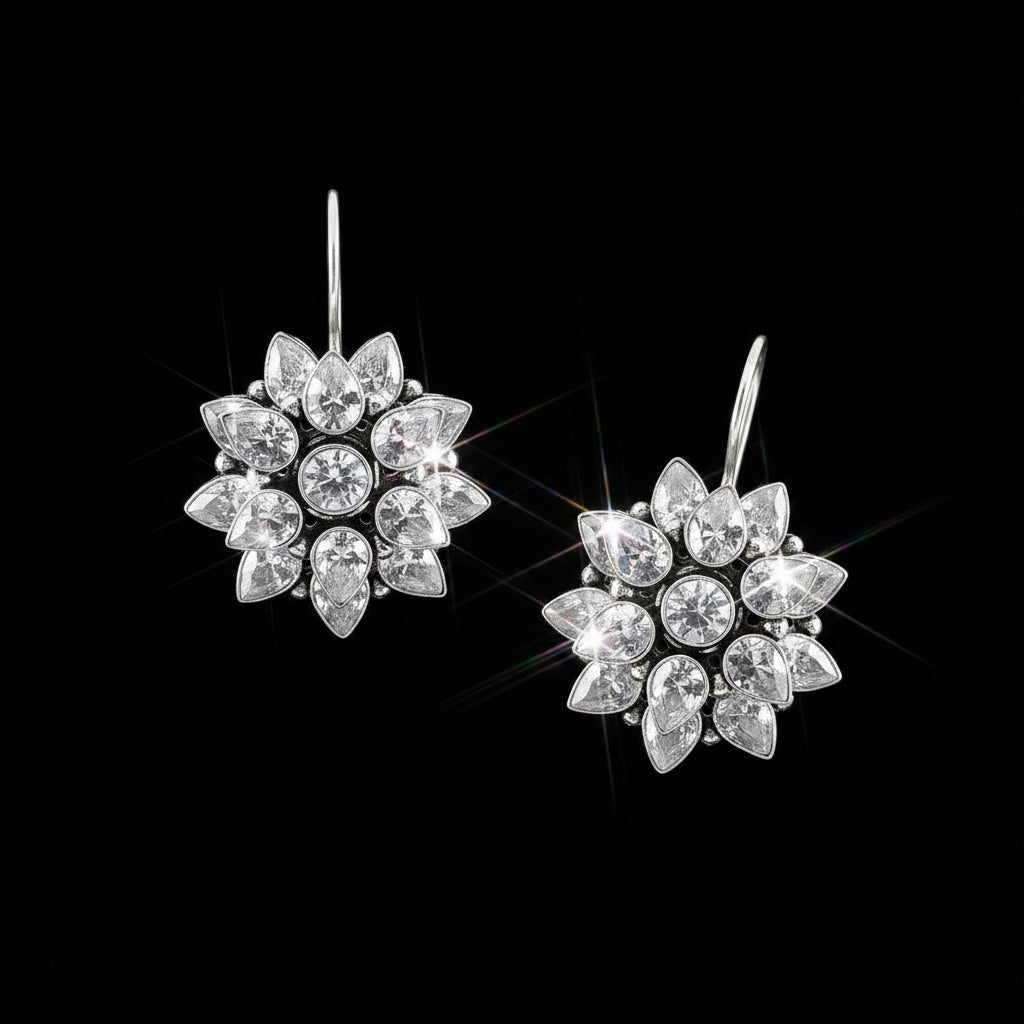 A pair of silver-toned dangle earrings with clear teardrop and round crystals arranged in a floral burst pattern, displayed on a reflective dark gray surface with the brand name SAHELI MY BEST FRIEND at the top.