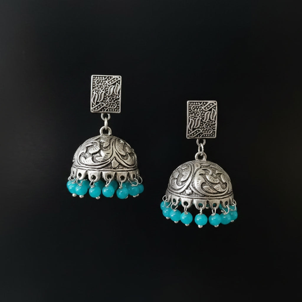A pair of traditional Indian silver jhumka earrings with turquoise blue bead drops, displayed on a gray velvet jewelry stand against a black background.