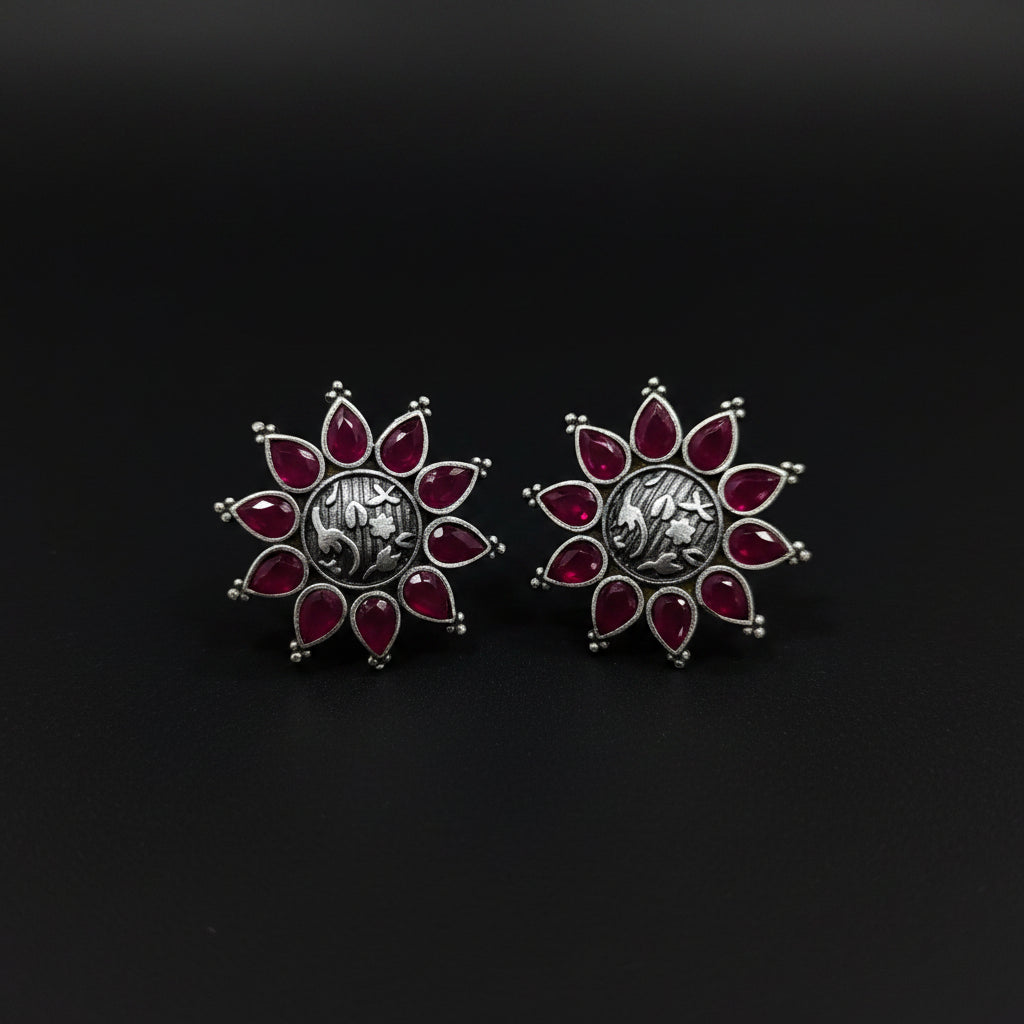 A pair of silver-toned floral stud earrings with deep red gemstone petals, displayed on a textured blue jewelry box with the brand name Al-Rawnaq visible.