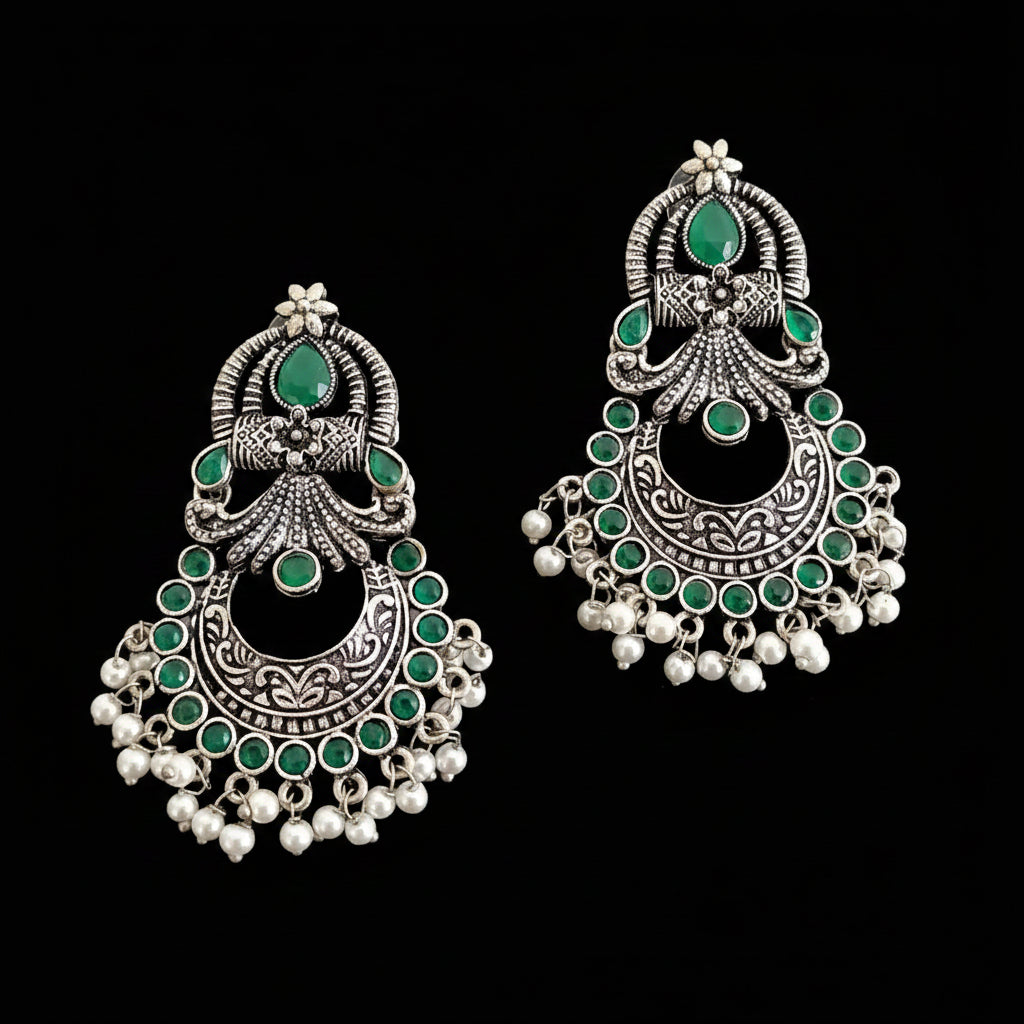 A pair of ornate silver-toned Indian jhumka earrings with green stones and white pearl-like bead drops, displayed on a dark reflective surface. The brand logo 'SAHELI BY SAHELI BRAND' is visible at the top center.