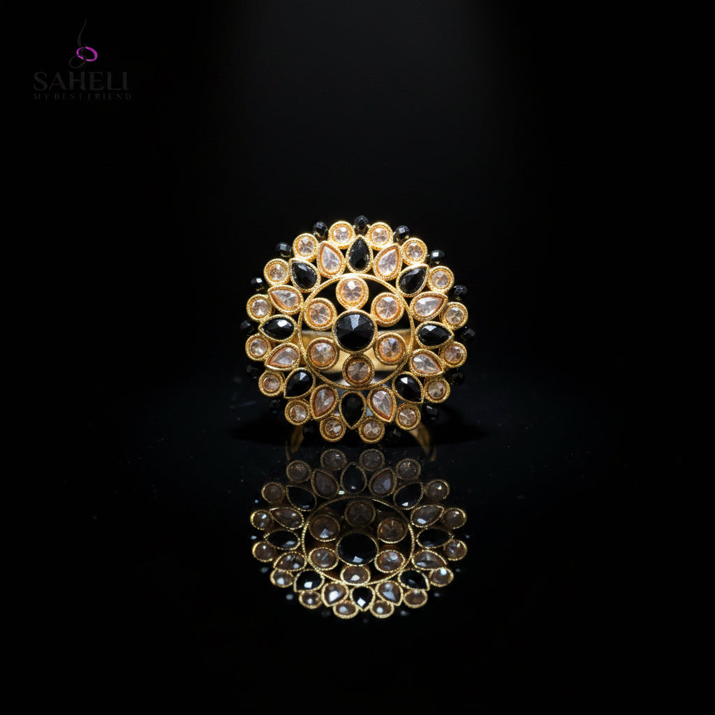 A gold-toned circular Kundan choker brooch featuring black stones and clear stones, displayed on a dark gray reflective surface with a Saheli My Best Friend logo in the top left corner.