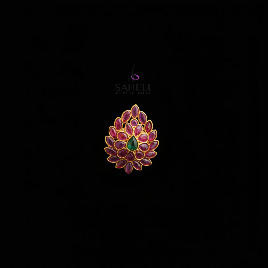 A gold ring featuring a floral mandala design with deep red enamel petals and a central green teardrop stone, displayed on a dark reflective surface with the SAHELI brand logo above.
