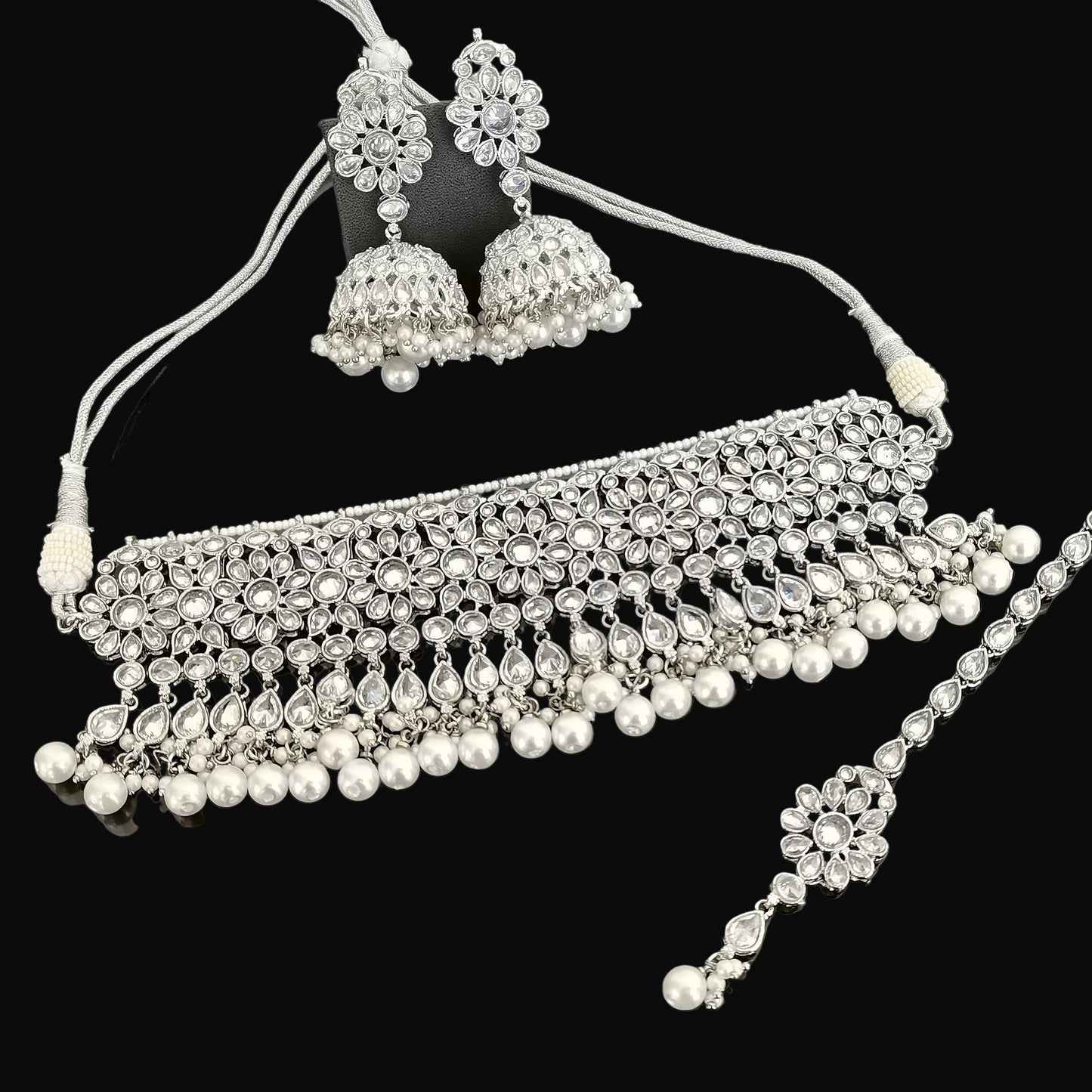 Silver jewelry set with necklace, earrings, and maang tikka on a dark surface with 'Saheli' branding.