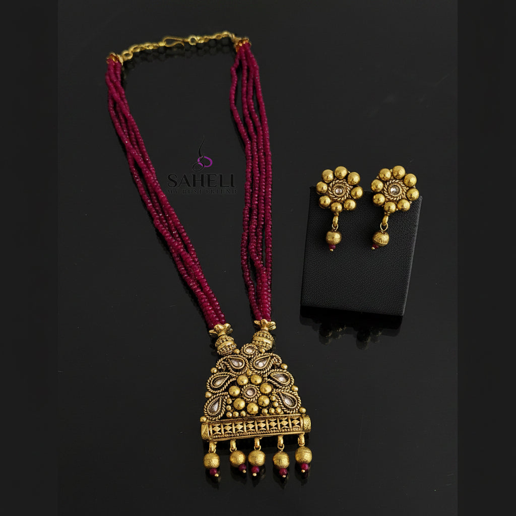 A traditional Indian jewelry set featuring a gold-plated pendant necklace with magenta pink beaded strands and matching gold flower-shaped stud earrings, displayed on a black reflective surface with a brand watermark 'SAHELI'.