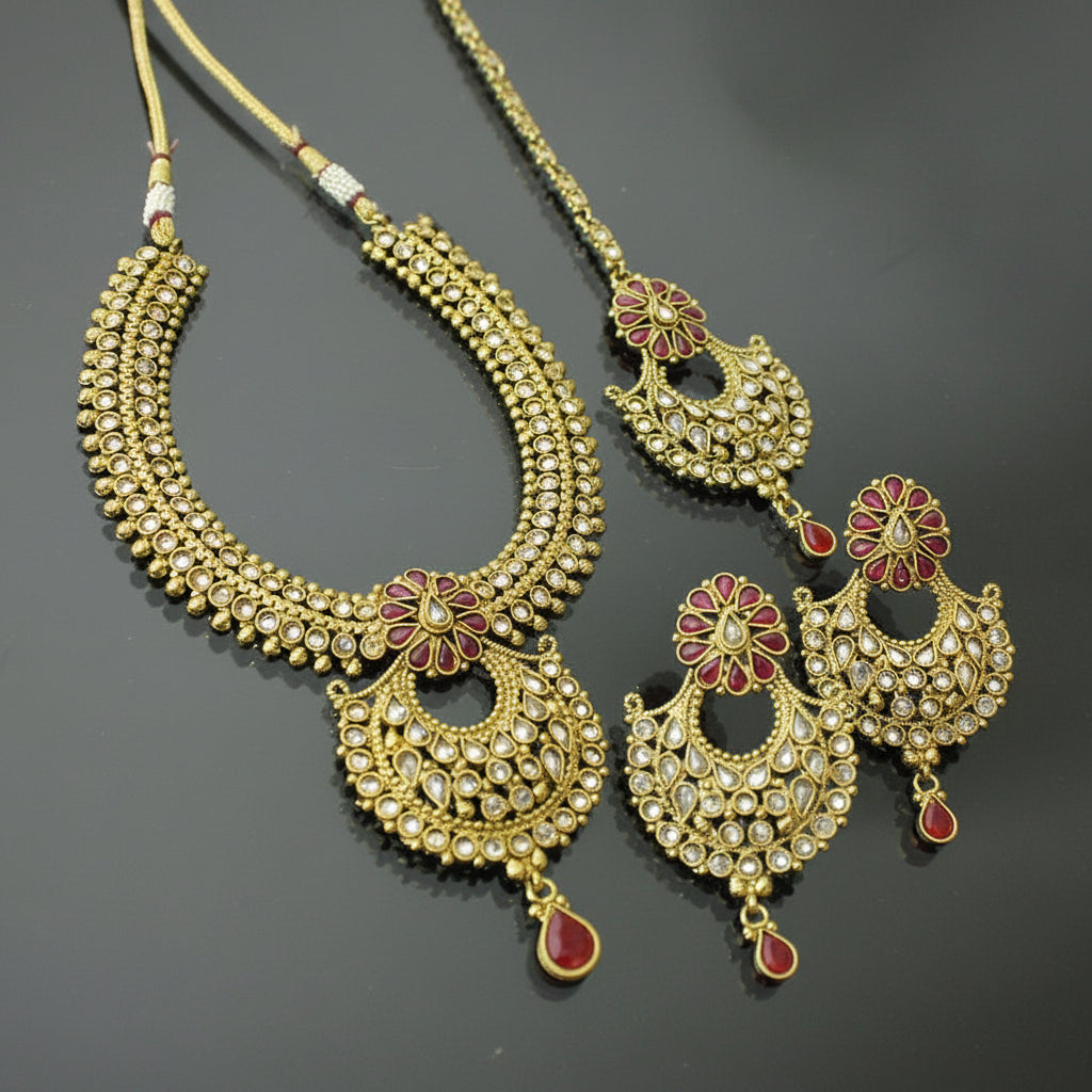 Gold-toned traditional Indian jewelry set featuring a necklace, earrings, and a matching pendant, embellished with clear stones and red floral accents, displayed on a dark reflective surface. The brand logo 'SAHELI MY BEST FRIEND' is visible in the top right.