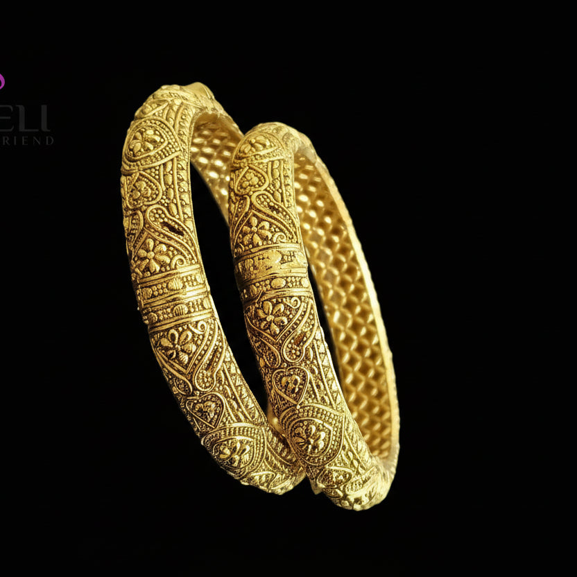 Pair of gold bangles with intricate designs on a reflective surface, featuring the brand 'Saheli'.