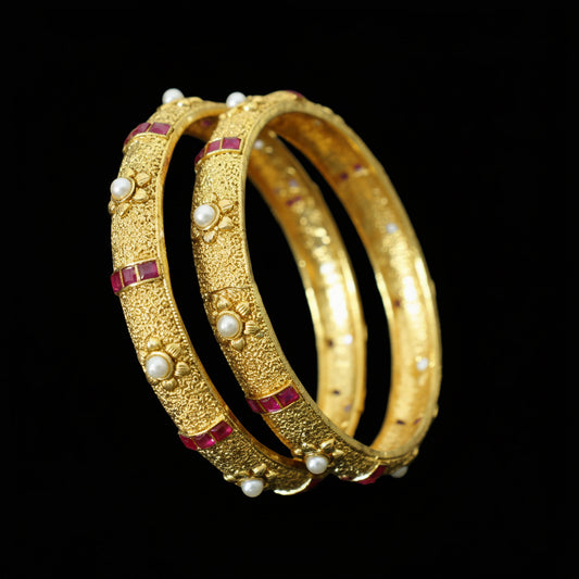 Gold bangles with pearls and red stones on a dark background, branded 'Saheli'.