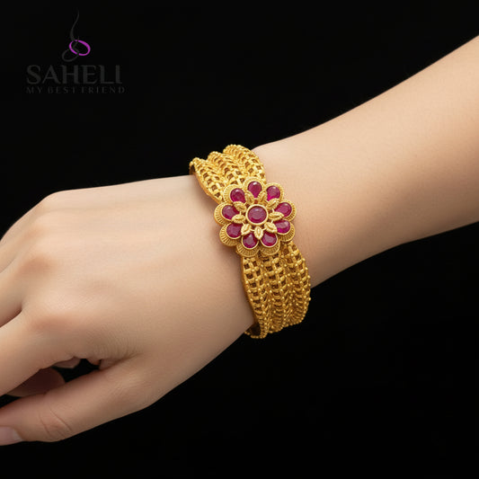 Gold bracelet with red stones on a reflective surface, branded 'SAHELI'.