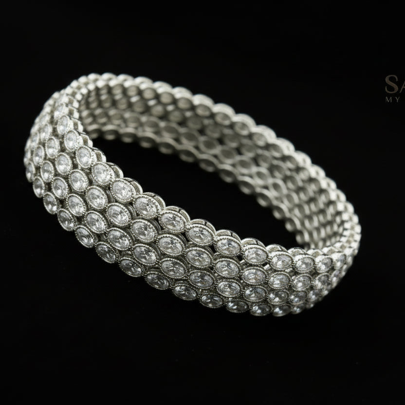 Silver bracelet with clear stones on a reflective surface, branded 'Saheli'.