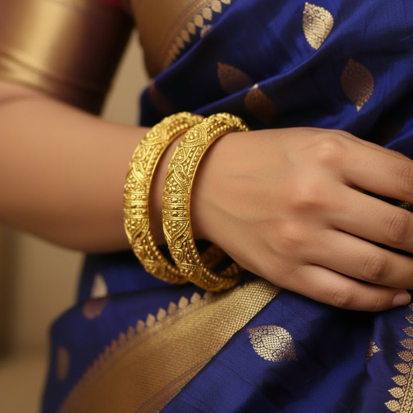 Two gold bangles with intricate designs on a reflective surface, featuring the brand 'Saheli'.