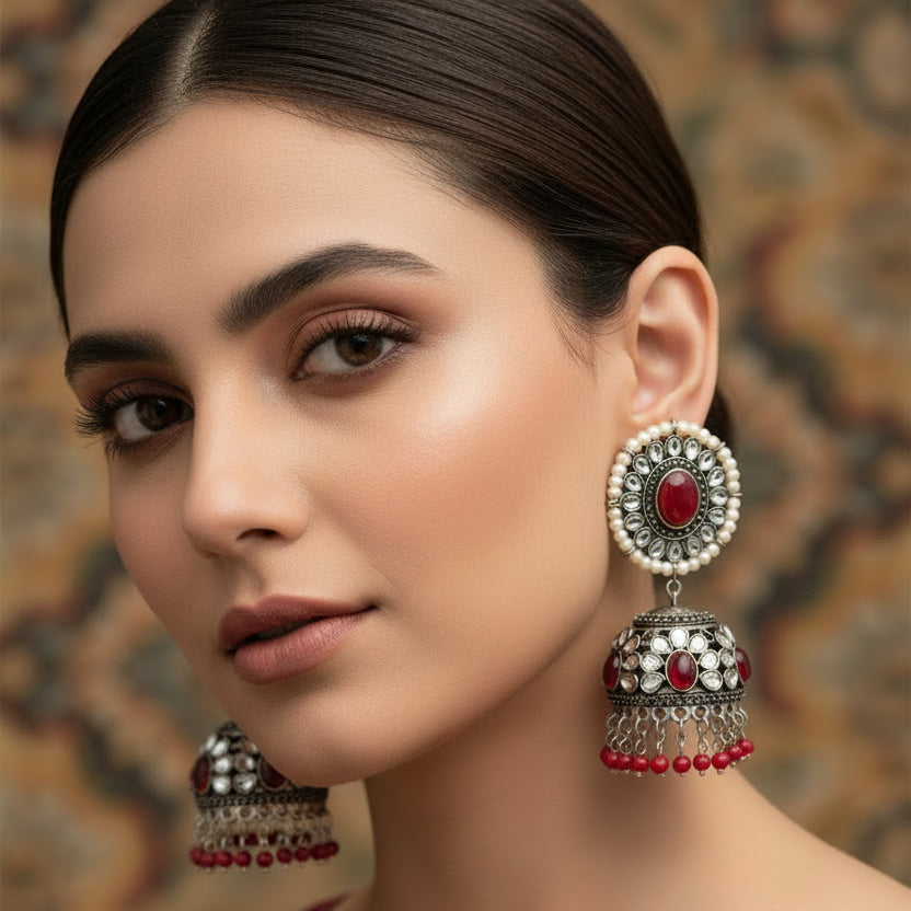 Pair of ornate earrings with red and silver details on a dark background, featuring the brand 'Saheli'.