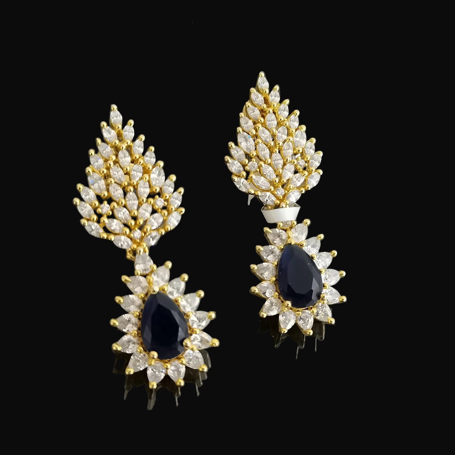Gold and diamond earrings with blue gemstones on a dark background, branded 'SAHELL'.