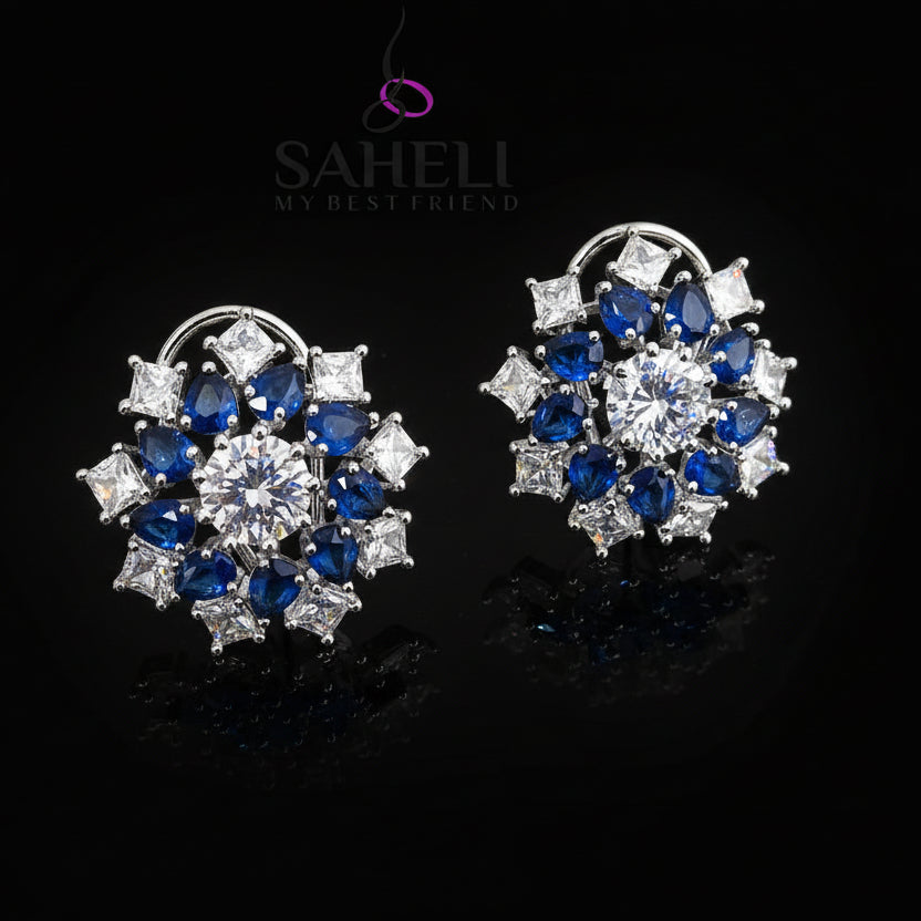 Pair of silver earrings with blue and clear gemstones on a gray background, branded 'Saheli'.