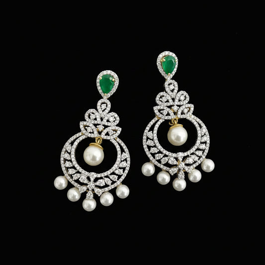 A pair of traditional Indian chandelier earrings by Saheli My Best Friend, featuring green stones, white pearls, and silver pavé settings on a dark reflective surface.