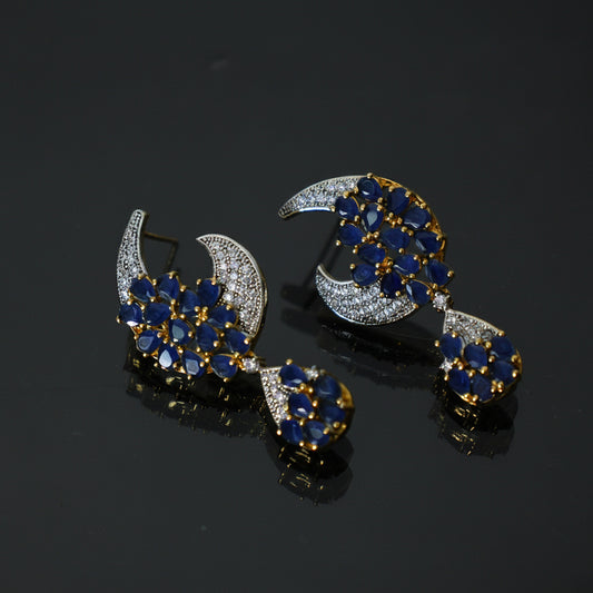 A pair of gold and silver crescent moon stud earrings decorated with blue and white stones, displayed on a dark reflective surface with a SAHELI MY BEST FRIEND logo in the upper left.