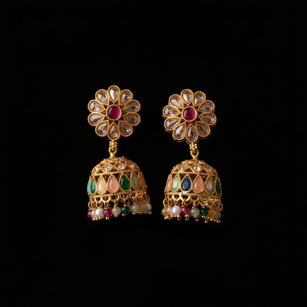 A pair of gold-plated Indian jhumka earrings with floral studs, clear stone accents, and colorful enamel details, displayed on a dark wood background.