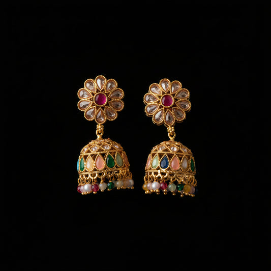 A pair of gold-plated Indian jhumka earrings with floral studs, clear stone accents, and colorful enamel details, displayed on a dark wood background.