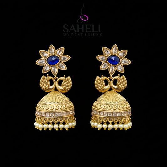 Gold jhumka earrings with blue gemstones on a gray stand, branded 'Saheli'.