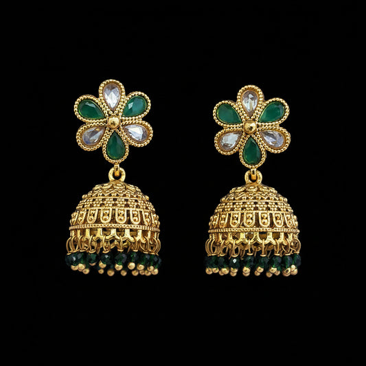 A pair of traditional Indian gold jhumka earrings with green stone floral tops and dangling bell-shaped charms, displayed on a textured blue background.