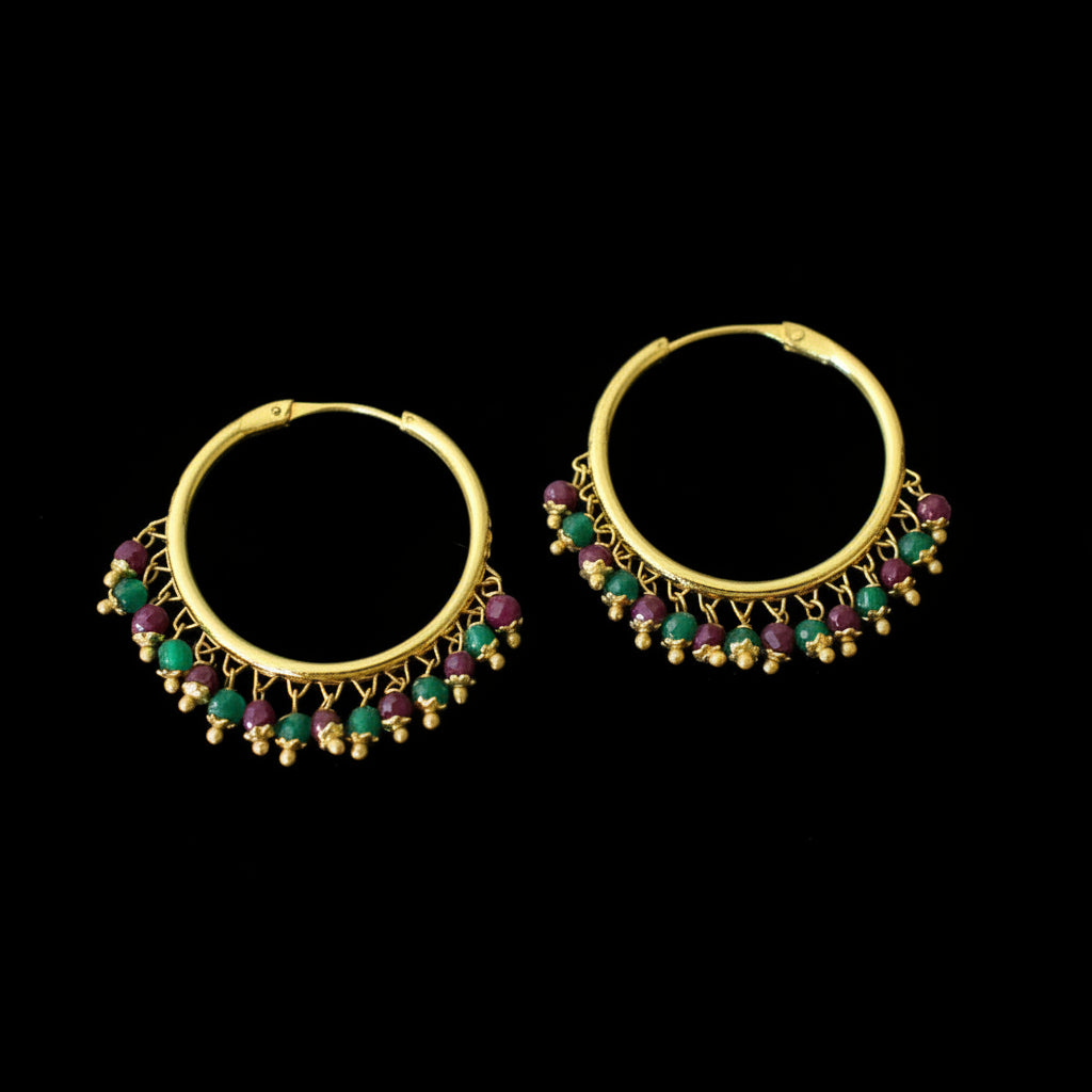 A pair of gold-plated hoop earrings with dangling chains of small gold beads and alternating green and pinkish-red teardrop gemstones, displayed on a dark reflective surface with the brand name 'SAHeli' at the top.