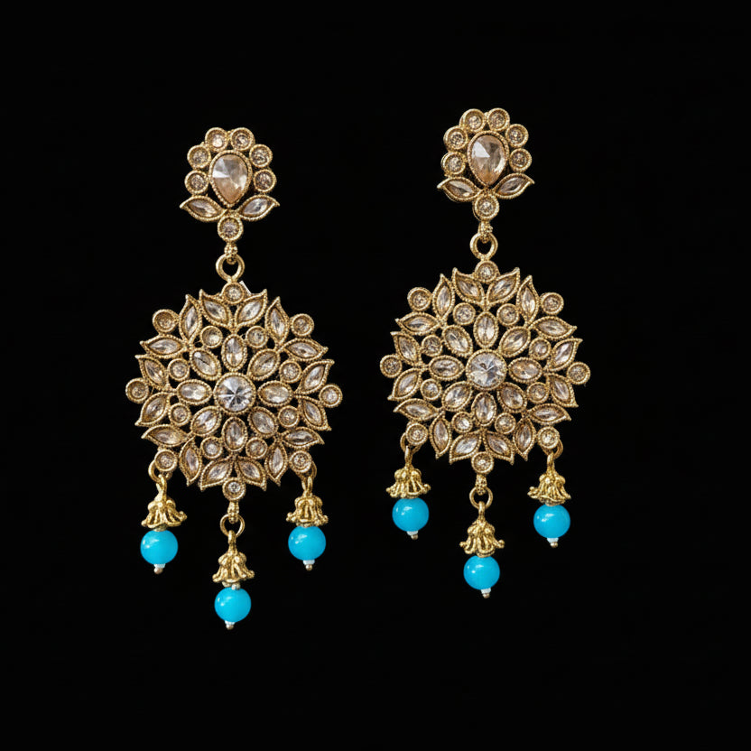 Ornate gold earrings with blue beads on a gray stand, featuring the brand 'Saheli'.