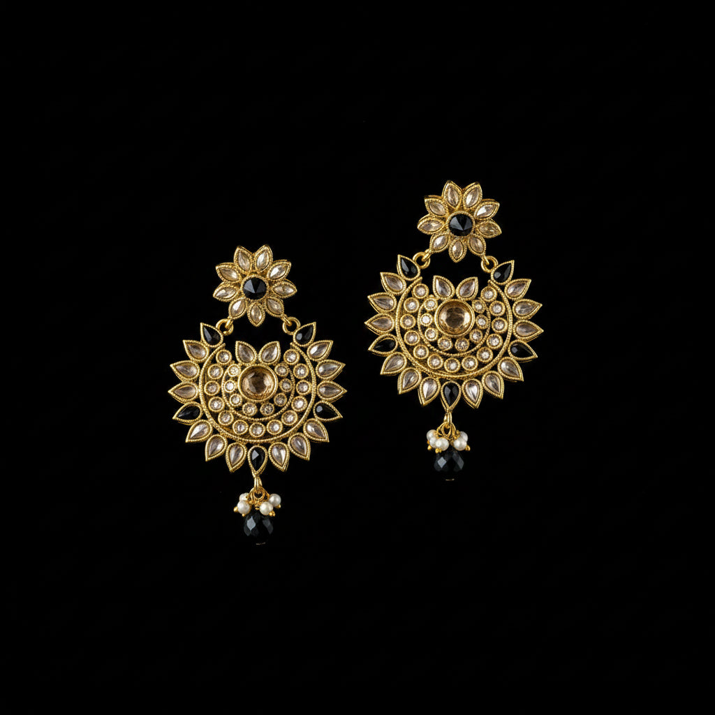 A pair of gold-toned Indian jhumka earrings with intricate Kundan and stone detailing, featuring black bead drops, displayed on a dark reflective surface with the Saheli brand logo above.