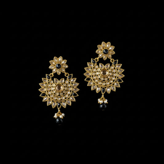 A pair of gold-toned Indian jhumka earrings with intricate Kundan and stone detailing, featuring black bead drops, displayed on a dark reflective surface with the Saheli brand logo above.