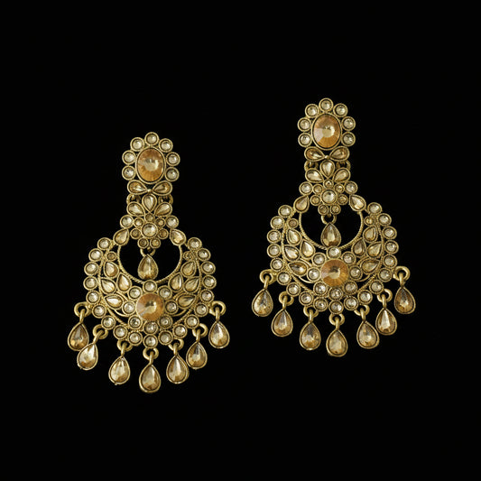 A pair of ornate gold-toned Indian chandbali earrings embellished with small white stones and teardrop-shaped gold drops, displayed on a dark reflective surface with the Saheli brand logo in the top left corner.