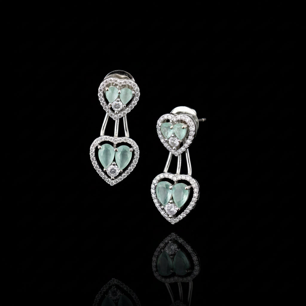 A pair of silver-toned drop earrings with heart-shaped mint green gemstones surrounded by small clear stones, displayed on a gray cushion.