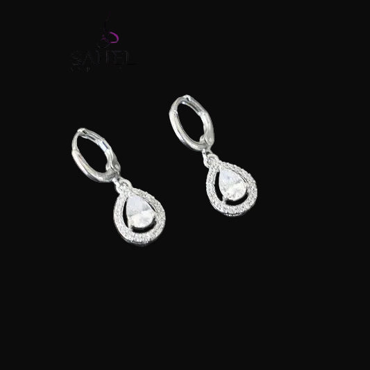 Teardrop-shaped silver earrings with clear stones on a dark background, branded 'SAHELI'.