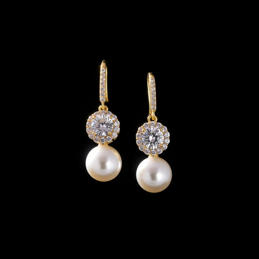 A pair of gold drop earrings with clear pavé-set round stones and a single white pearl dangling below, displayed on a teal background with decorative stones.