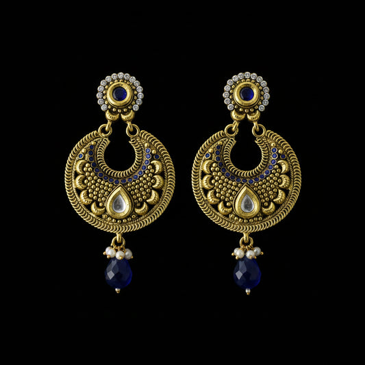 A pair of traditional Indian gold-toned dangle earrings with intricate filigree, white teardrop inlays, small clear stones, and dark blue bead drops, displayed on a dark reflective surface.