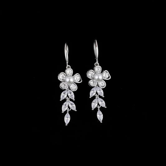 A pair of silver-tone dangling earrings with floral and leaf motifs set with clear rhinestones and a central pearl, displayed on a teal background with decorative stones and greenery in the corner.